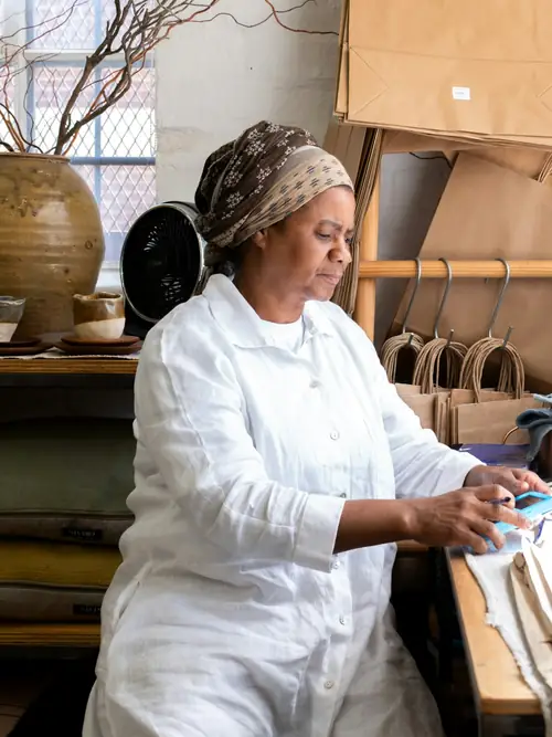 A woman working at her bench in a small workshop, paper carriers on the rack behind her